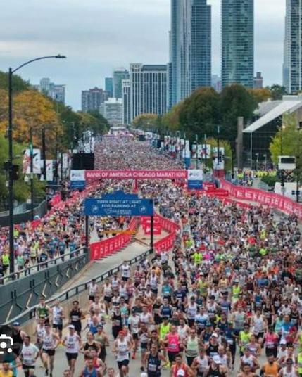 Thousands of runners fill the streets of downtown Chicago at the start of the Bank of America Chicago Marathon. A large red and blue banner marks the starting line, surrounded by cheering crowds, colorful race bibs, and city skyscrapers in the background under clear skies.