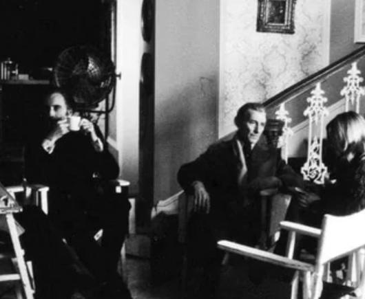 A black-and-white photograph of best friends and British actors Christopher Lee and Peter Cushing on the set of the 1973, British, Hammer Horror film “The Satanic Rites of Dracula”. They are both chatting with film crew members while drinking tea and wearing suits.