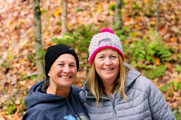 Photo of the head and shoulders of two women,  both wearing hoodies,  one a puffer and caps with fallen leaves in the background