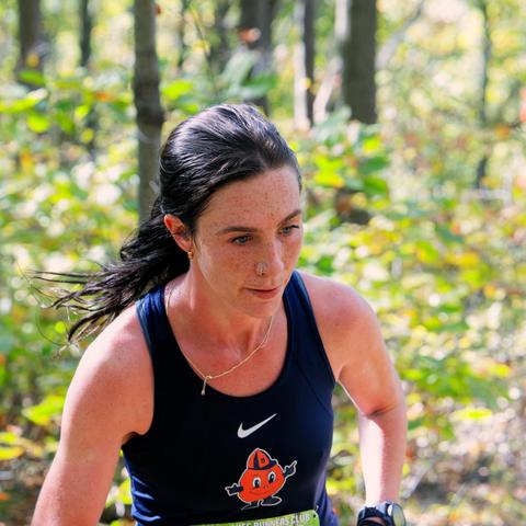 A woman in the middle of a square frame wearing a syracuse orangemen tank top with a nike logo, a necklace and with her dark hair tied back.  In the background some green and yellow backlit understory vegetation
