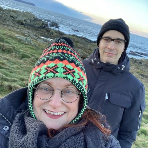An outdoor selfie-style photo of a woman and man standing along a rocky, northern ocean coastline. Both are wearing eyeglasses, winter coats and knit hats; both are smiling and appear happy. A partially cloudy sky can be seen in the background.