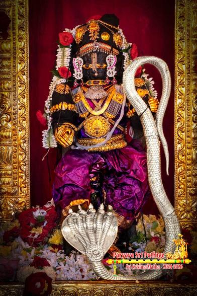 Closeup of idol of Hindu deity Krishna in bright purple holding a 5-headed silver serpent by its tail in left hand and appearing to be stomping/dancing on the heads of the serpent (though his feet are not visible). Krishna wears gold palm coverings, lots of gold ornaments, and a string of white flowers (some red roses) waist-waist. The sides of a gold arch behind Krishna are visible. Green holy basil leaves and assorted loose flowers at Krishna's feet.