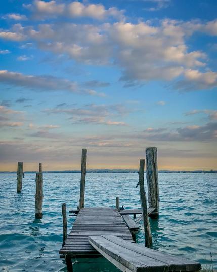 Old wooden pier with missing planks extending into turquoise-blue waters of Lake Garda near Sirmione, Italy, with scattered clouds glowing in the warm light of sunset and distant shoreline in the background.