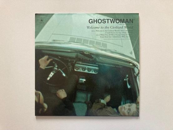 Albumcover für "Ghostwoman: Welcome to the Civilized World". Das Bild zeigt eine Draufsicht auf zwei Personen in einem Auto, wobei der Schwerpunkt auf den Händen des Fahrers am Lenkrad liegt. Im Hintergrund ist eine verschwommene Straßenszene zu sehen.