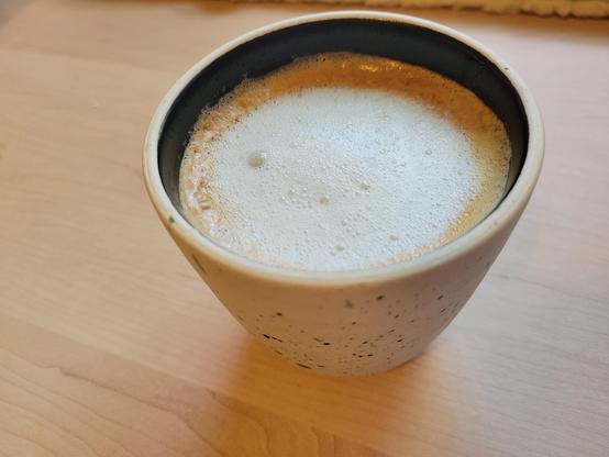 12-ounce, handle-less, off-white ceramic cup with a coffee latte, on a maple desk surface. The clay of the cup has many tiny flecks of black and blue. The interior of the cup has a dark-colored glaze. 