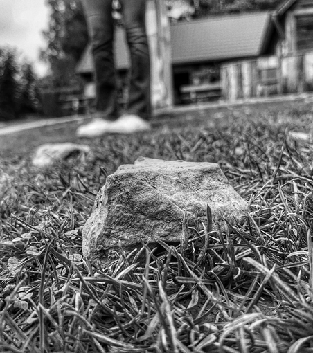 Black and white image of stone in foreground and person’s legs and feet blurred in background 