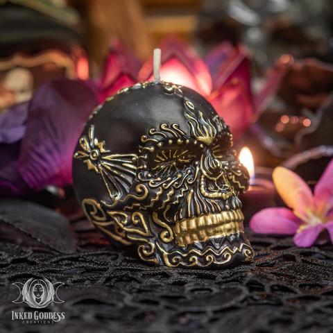 An example of the Black & Gold Skull Candle sits on a black lace-covered surface. This small black candle is shaped like a human skull and is covered in embossed gold designs. Presented by Inked Goddess Creations.