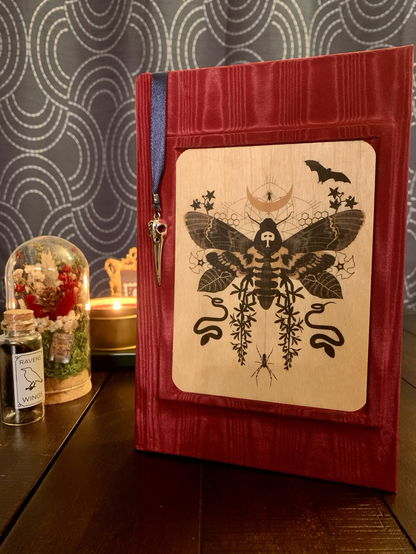 A handmade journal is standing upright on a dark wooden table. The bookcloth is a metallic red color, and glued and centered on the front cover is a wood veneer card with an illustration of a moth surrounded by spooky motifs (a spider, snakes, a moon, bats, and plants). The book has a blue ribbon bookmark hanging from the top edge of the book. Attached to the end of the bookmark ribbon is a silver colored metal charm in the shape of a bird skull. In the background around the book is a small glass cloche containing dried flowers, a candle in a bronze colored tin, and a small corked glass vial with black feathers inside. The vial is labeled “Raven feathers”. 