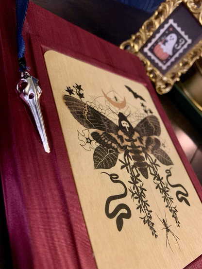 The photo is zoomed in on front cover of the red journal to show the details of the wood veneer illustration of the moth surrounded by flowers, bats, snakes, and a moon. The silver bird skull at the end of the blue ribbon bookmark is also visible, hanging from the top edge of the book.