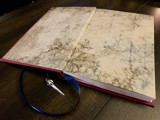 The red journal is open to show the front end paper of the book. The end paper is a handmade paper designed to look old, stained, and weathered. 