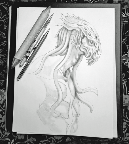 A black and white pencil sketch by Kmac of a creature with a large, multi-eyed head and flowing tentacle-like appendages, with two pencils and one blender stump lying nearby. 
The head features a ridged, elongated appearance with multiple small eye-like markings and a toothy mouth, while the body is partially sketched with tendrils draping downward. The paper is resting on an illustration portfolio with an unfinished quilt top background.