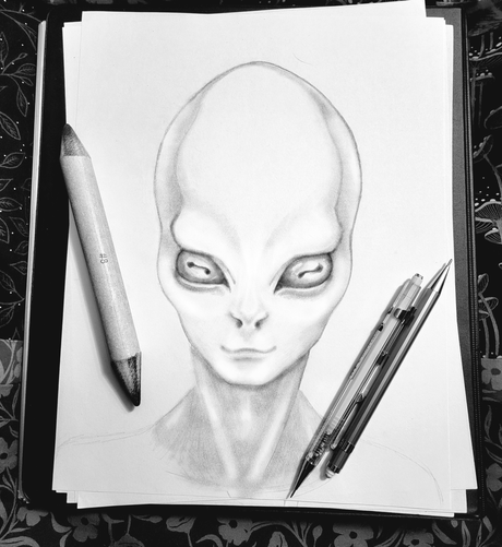 A  pencil sketch by Kmac of an alien with a large, oval head, almond-shaped eyes, and a subtle smile, resting on a stack of sketching paper with two pencils and a blending stump. All sitting atop an illustration portfolio with an unfinished quilt in the background.

