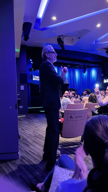Jeff Goldblum enters the Recording Academy theatre a suit stands on stage, addressing an audience in a brightly lit venue. The background features blue lighting and musical instruments, hinting at a live performance or event. Audience members are seen clapping and engaging with the presentation.
