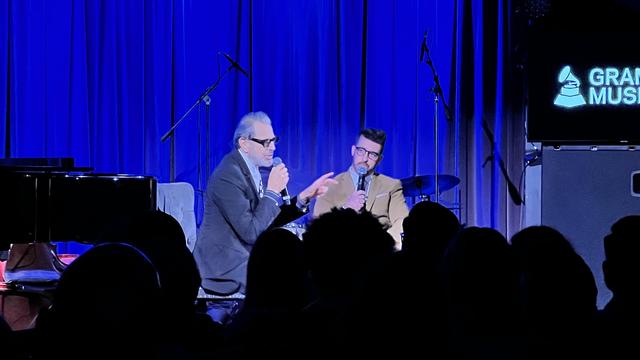 An event scene featuring two speakers in conversation on stage, illuminated by blue lighting. Jeff holds a microphone and gestures, while the host Paul Costabile listens attentively. In the background, a grand piano is visible and a sign reading "GRAMMY MUSEUM.”