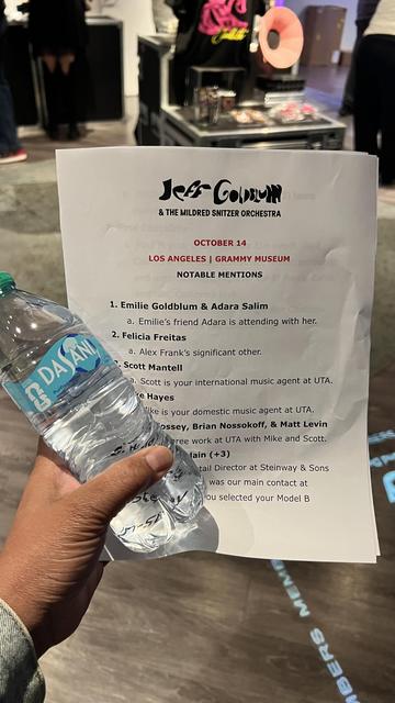 A hand is holding a bottle of Dasani water and a printed sheet detailing notable mentions for an event featuring Jeff Goldblum and the Mildred Snitzer Orchestra at the Grammy Museum in Los Angeles. The background includes people and promotional items.