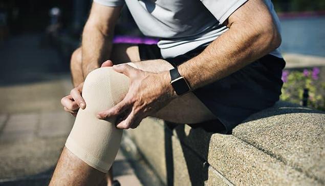 A person wearing a knee support brace while sitting outdoors, gently holding their knee to relieve pain — representing knee pain management, joint support, and mobility care.