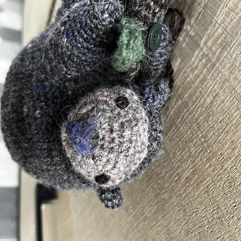 Close up of an amigurumi sloth hanging from a crocheted branch with a leaf visible. The whole thing is attached to a wooden kitchen cabinet. The body is in a tonal darker grey and the face in a pinkish light color. The nose is felted in brown with some purple and the eyes are french knots in brown. All of the yarn is hand spun.