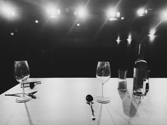 S/W-Aufnahme von einer Bühne in Richtung Publikumsbereich. Auf einem Tisch auf der Bühne stehen zwei Weingläser, Löffel, eine Weinflasche und Salz- und Pfefferstreuer. Das Auditorium ist aufgrund der sechs hellen Scheinwerfer, die die Bühne beleuchten, kaum bis gar nicht zu sehen. 