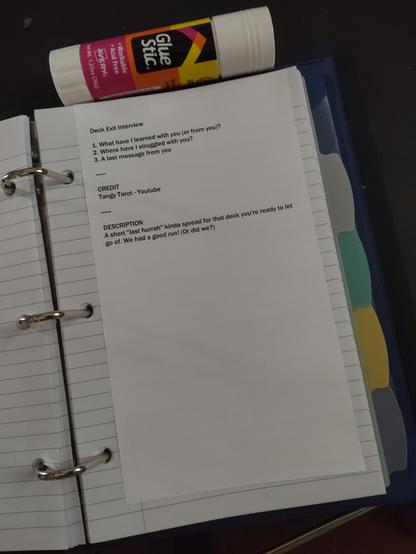a mini binder laying open. I printed a tarot deck exit interview and cut the paper to fit inside the binder. the printed sheet has been glued to a three hole punched lined paper inside the binder. a glue stick sits above the binder. there are five divider panels behind the sheet.