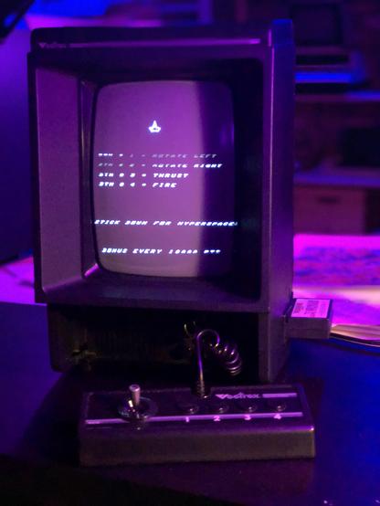 A black monitor console gaming unit with a portrait oriented display and a joystick in front. The lighting is giving things a magenta hue. 