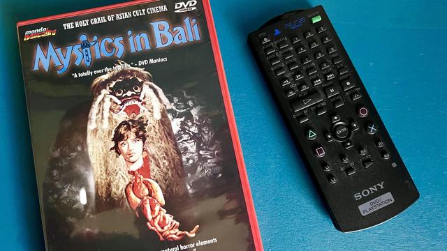 DVD box of “Mystics in Bali” beside a PlayStation 2 remote control. Both sit atop a blue end table. 