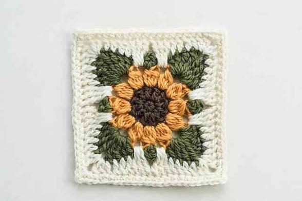 Square crocheted granny square featuring a central sunflower design with dark brown, orange petals, and olive green leaves, framed by white yarn against a light background.