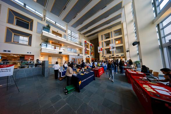People bill around numerous tables set out to represent various educational institutions in a tall atrium flanked by windows on the right and two other engineering buildings on the right and back