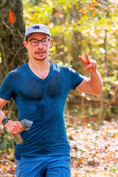 Male runner with a blue shirt and blue pants and billed cap has a short beard, holds a crumpled bottled in his right (your left) hand which has a watch on that wrist and makes a v sign with the fingers of his other hand in a fall color forest