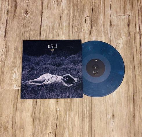 Ein Albumcover mit dem Titel "KALI", das ein Schwarz-Weiß-Bild einer im Gras liegenden Frau neben einer blauen Schallplatte zeigt. Der Hintergrund ist mit einer Holzbodenstruktur versehen.