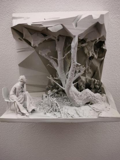 Biała rzeźba przedstawiająca siedzącą na krześle postać kobiecą w długiej sukni, pogrążoną w zadumie przed surrealistycznym krajobrazem. W centrum kompozycji wyrasta drzewo o chropowatej fakturze i nierealnych kształtach, otoczone fragmentami konstrukcji przypominających ruiny lub szczątki. Całość ma intymny, melancholijny nastrój i przypomina senne, oniryczne wspomnienie natury.