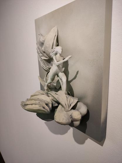 Trójwymiarowa rzeźba w jasnych, matowych tonach przedstawiająca postać kobiecą w ruchu, jakby wychodzącą z powierzchni ściany. Figura otoczona jest formami przypominającymi liście lub organiczne płaty, które tworzą dynamiczne, falujące tło. Kompozycja sprawia wrażenie połączenia natury i ciała w jednym płynnym, ekspresyjnym geście.