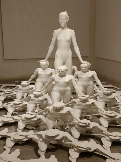 Instalacja rzeźbiarska przedstawiająca wiele białych, gładkich i stylizowanych nagich postaci.
Na szczycie stoi wysoka, smukła postać kobieca o spokojnym wyrazie twarzy, której dłonie spoczywają na ramionach trzech figur poniżej. Te trzy postacie to również nagie kobiety, zwrócone przodem, z głowami lekko pochylonymi w dół.
Poniżej i wokół nich znajduje się wiele podobnych, mniejszych nagich postaci, niektóre wydają się być męskie (ze względu na krótsze fryzury przypominające czapki lub kapelusze), wyłaniających się z chaotycznej, warstwowej podstawy z płaskich, białych, nieregularnych kształtów pokrywających podłogę. Ogólne wrażenie to duża grupa figur stapiających się z fragmentarycznym, płaskim krajobrazem, wszystko w monochromatycznej bieli.