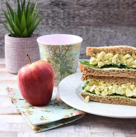 Vegan egg mayo sandwich on soft brown bread stacked on a plate next to an apple & cup of water.