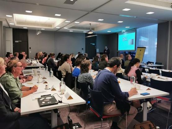 Participants assis en rangées, prenant des notes lors d’une session plénière animée par un intervenant, dans une salle de conférence équipée de projecteurs.