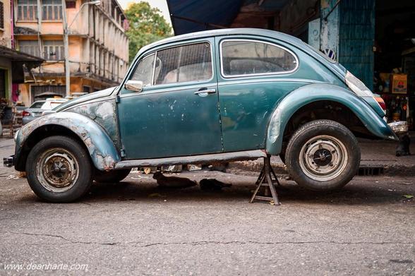 Side view of a teal 1970s Volkswagen Beetle 1300 on jack stands in a Bangkok alley garage, showing its flaking paint, rust, and classic curved design.
