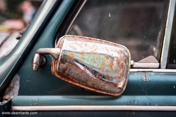 Detail of a rusty chrome side mirror on a teal 1970s Volkswagen Beetle 1300, with textured patina and reflections of the street in Bangkok.