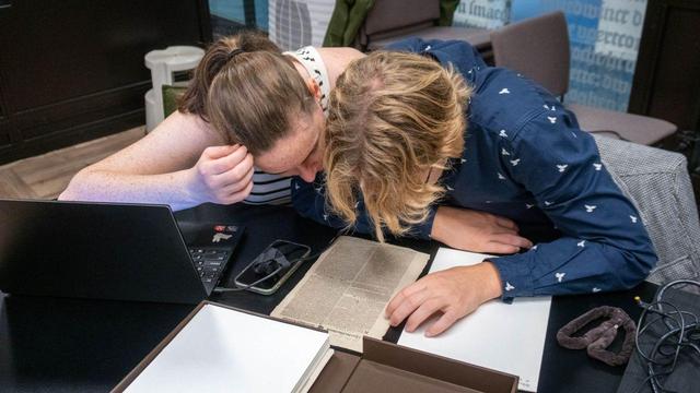 Twee studenten naast een laptop buigen zich over een oude krant, een dicht bedrukt vel papier op ongeveer A4-formaat.