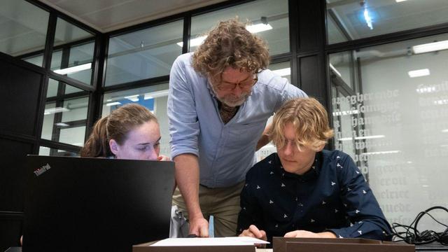 Een man buigt zich over twee studenten aan een bureau.