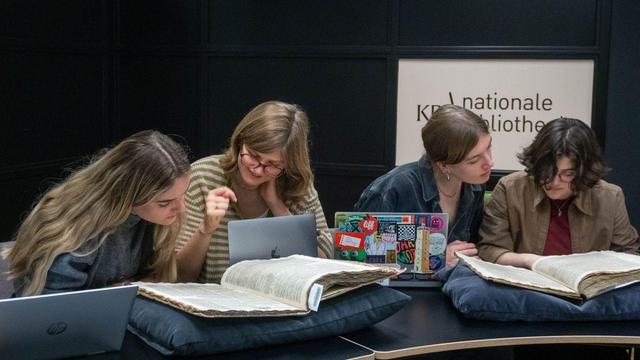 Vier studentes met laptops op een rij kijken naar twee grote banden met ingebonden oude kranten. 