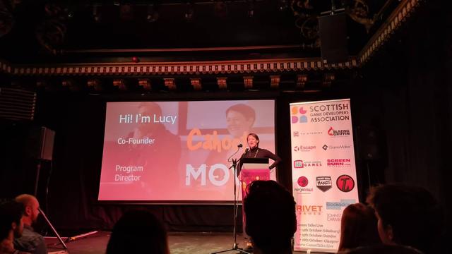 Pic of stage at Games talks live, screen front centre, overhead ornate scrollwork on ceiling, speaker and lectern to picture right, white popup banner with sponsors logos to the far right, orangey reddish lighting