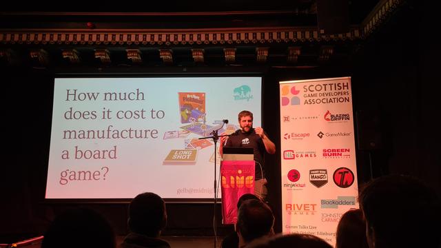 Pic of stage at Games talks live, screen front centre, overhead ornate scrollwork on ceiling, speaker and lectern to picture right, white popup banner with sponsors logos to the far right, orangey reddish lighting