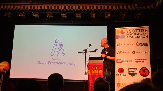 Pic of stage at Games talks live, screen front centre, overhead ornate scrollwork on ceiling, speaker and lectern to picture right, white popup banner with sponsors logos to the far right, orangey reddish lighting