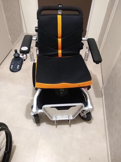 Photo d'un fauteuil roulant électrique dont le dossier est noir avec une bande orange au milieu. Le coussin d'assise est également noir avec les bords oranges. Le cale pied monobloc est remonté.