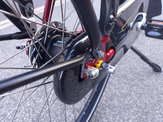 This close-up image focuses on the rear wheel and braking system of a bicycle. The wheel has a black tire and numerous silver spokes radiating from a central hub. A silver chain tensioner is prominently displayed, attached to the axle. The chain is completely enclosed in a shiny black chain case. A silver roller brake radiator is visible left of the 7-speed Nexus gear hub.
