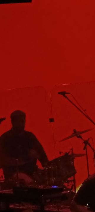 Silueta negra a contraluz de un hombre en una batería, tocando. El fondo es rojo. Se ven las partes de la batería y algunos micrófonos.