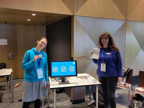 two pyladies standing next to their conference booth, holding up some merch