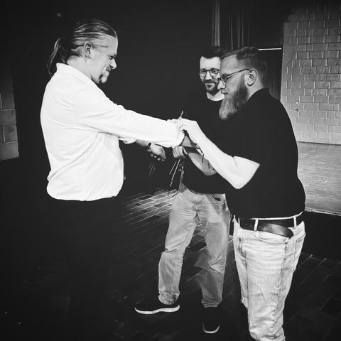 S/W-Fotos von zwei Männern, die sich vor einer Bühne gegenüberstehen, ein dritter im Hintergrund beobachtet lächelnd die beiden. Der rechte der Männer krempelt dem linken den Ärmel seines weißen Hemds hoch. 