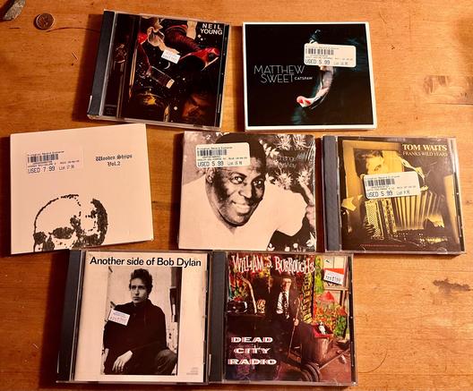 7 used CDs: (starting top-left) Neil Young, American Stars ‘n’ Bars; Matthew Sweet, Catspaw; Wooden Shjips, Vol. 2; Howlin’ Wolf, Change My Way; Tom Waits, Franks Wild Years; Bob Dylan, Another Side; & William S. Burroughs, Dead City Radio. 
