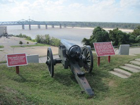 Historic cannon mounted on a grassy bluff overlooking the Mississippi River in Vicksburg, Mississippi. The cannon faces outward toward the river and bridge in the distance, flanked by two red interpretive signs describing the site’s Civil War history. The view emphasizes the city’s strategic position during the 1863 siege.