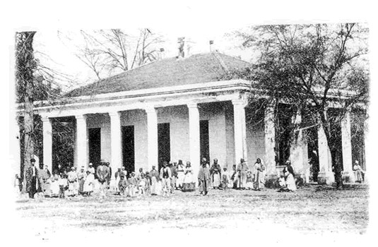 Black-and-white photograph of a plantation house with tall white columns and a hipped roof, surrounded by a large group of African American men, women, and children—likely formerly enslaved people—standing in front of the building. The scene captures the main house at Davis Bend, Mississippi, once part of the plantation owned by Joseph Davis, brother of Jefferson Davis.